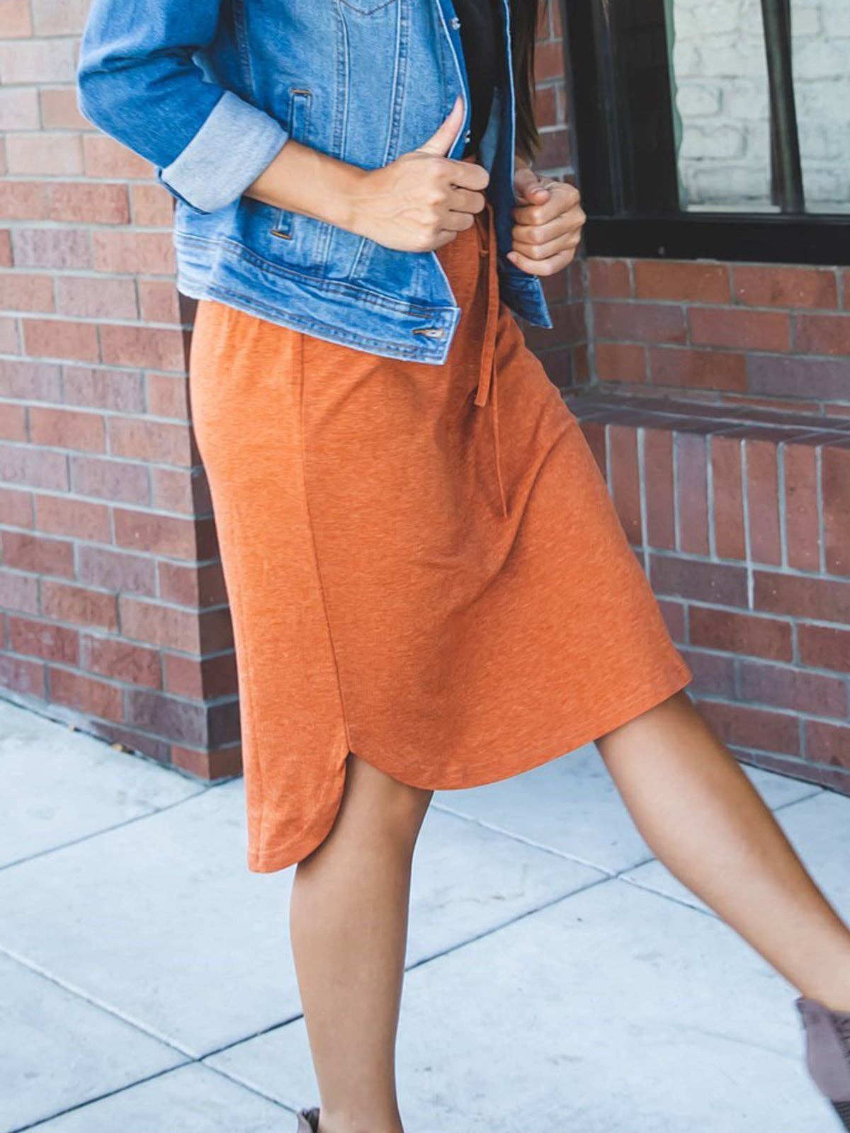 Person wearing an orange skirt and blue denim jacket in front of a brick wall.