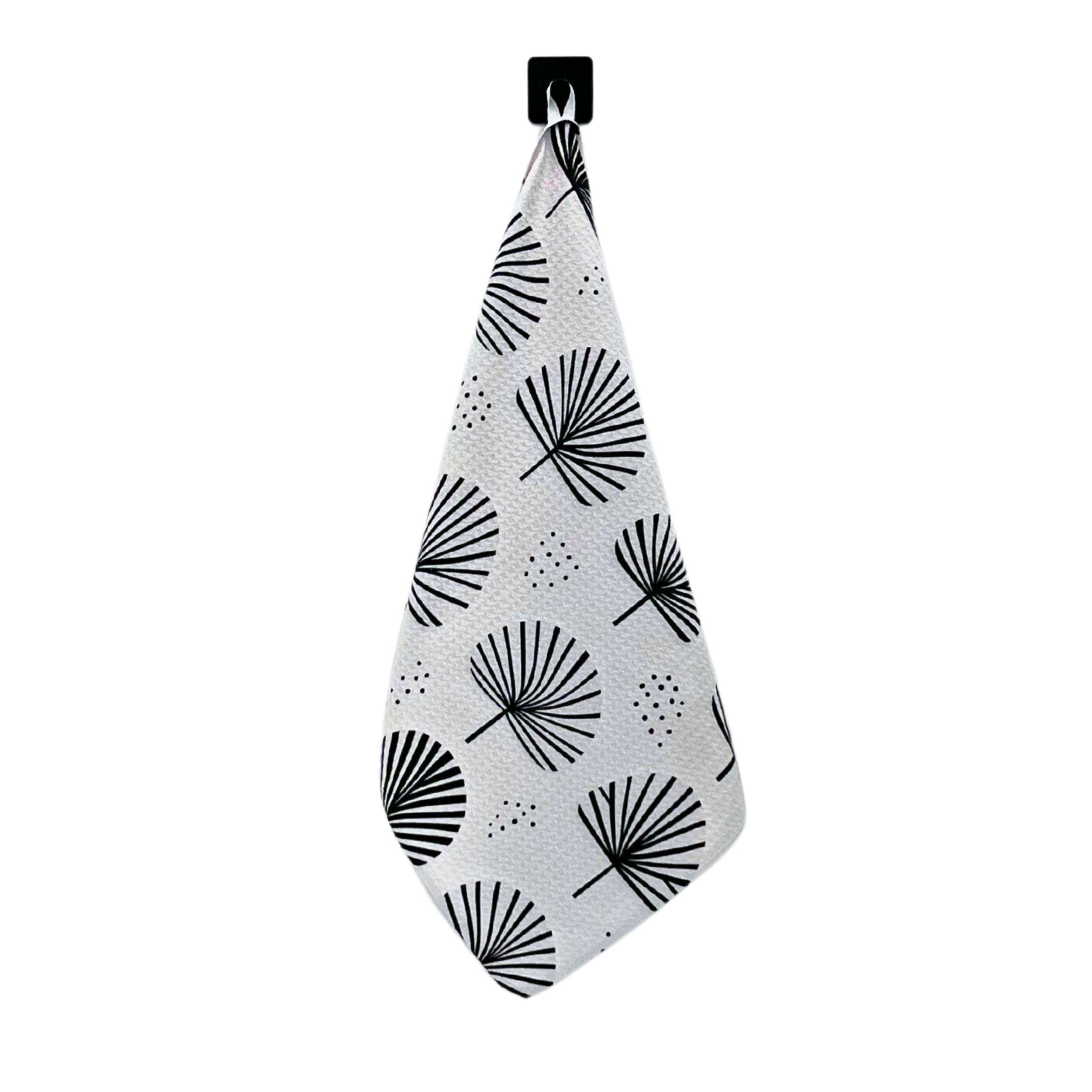 A white kitchen towel with a black palm frond pattern, hanging from a black wall hook against a solid white background.