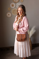 Woman in a pink sweater and white skirt holding a brown leather bag in a room with decorative wall art.