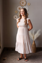 Woman in a tan dress standing in a room with decorative wall art and a vase.