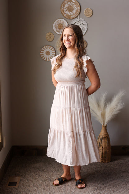 Woman in a tan dress standing in a room with decorative wall art and a vase.