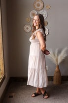 Woman in a tan dress standing in a room with decorative wall art and a vase.