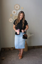 Woman wearing a black button up blouse and light wash denim skirt, holding a drink and a bag, standing in a room with decorative wall art.