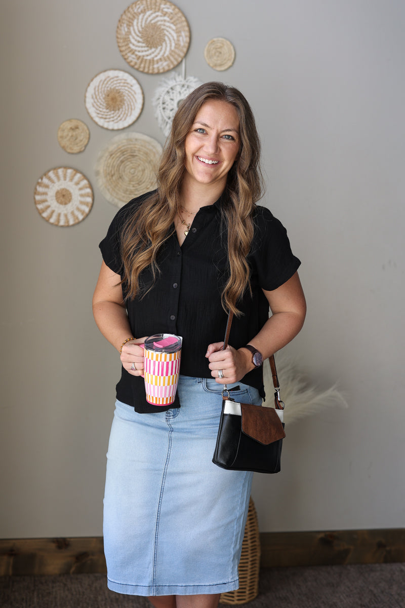 Woman wearing a black button up blouse and light wash jean skirt holding a colorful cup and a black handbag in a room with decorative wall art.