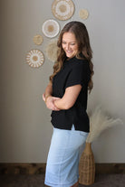 Woman in black top and light blue jean skirt standing in a room with decorative wall art.