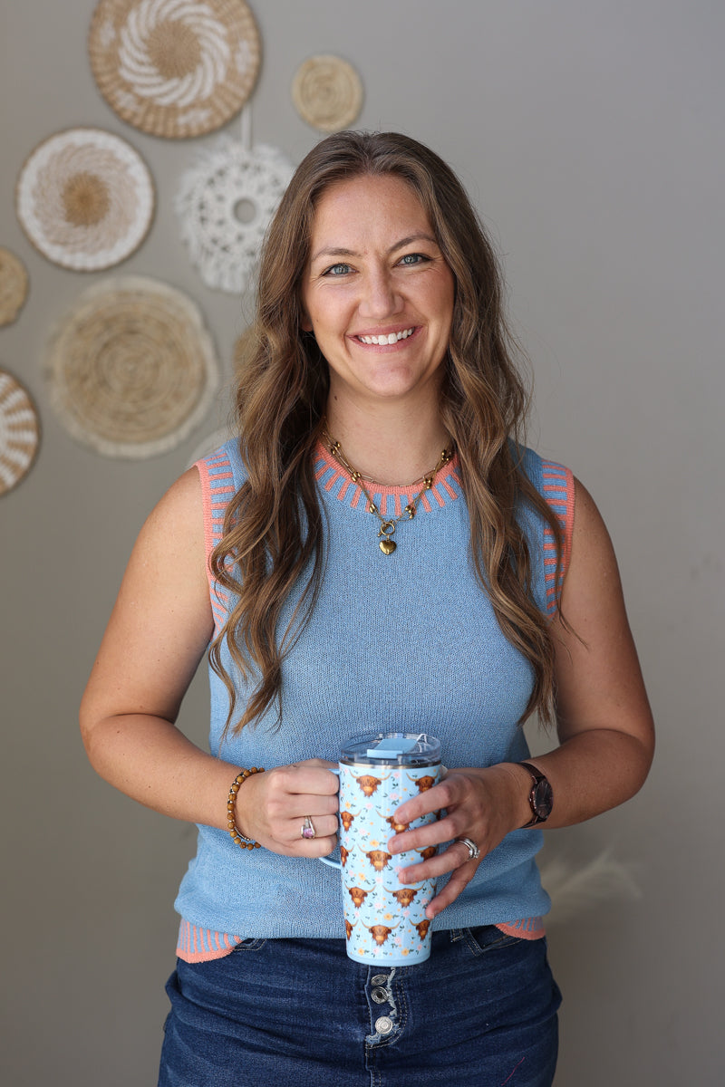 Woman wearing a blue sleeveless sweater with coral contrast, holding a patterned tumbler against a neutral wall with decorative plates.