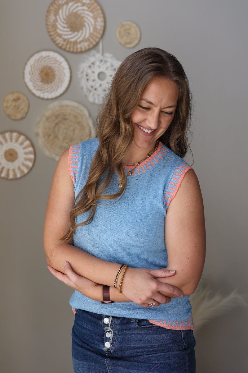 Woman wearing a blue sleeveless top with decorative trim, standing against a wall with decorative plates.