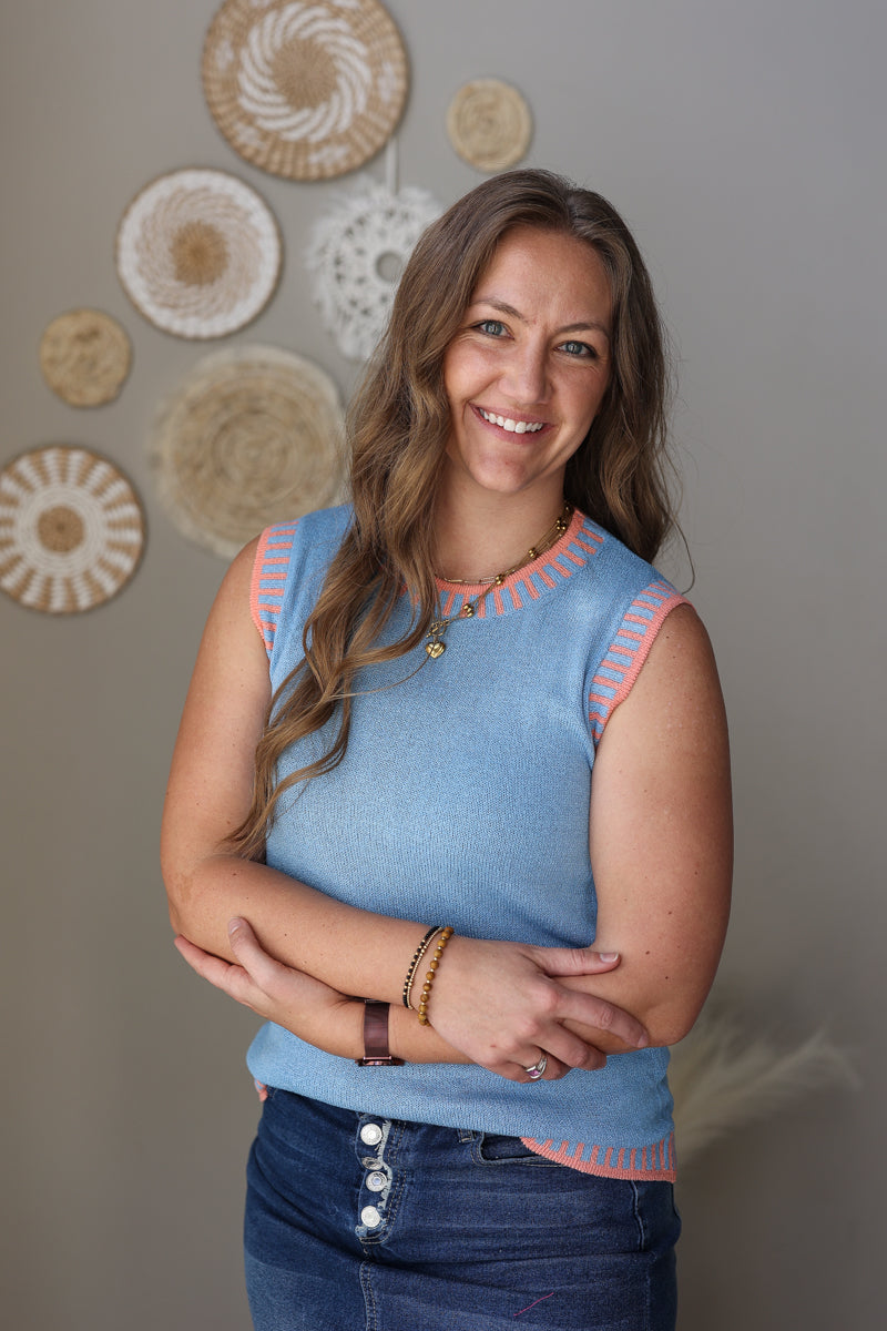 Woman wearing a blue sleeveless top with pink trim, standing against a wall with decorative circular patterns.