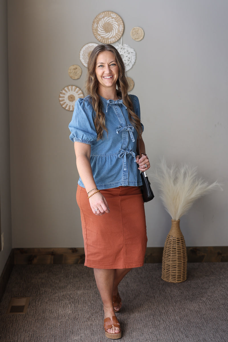 Woman in a blue top and rust-colored skirt standing in a room with decorative wall art and a vase.