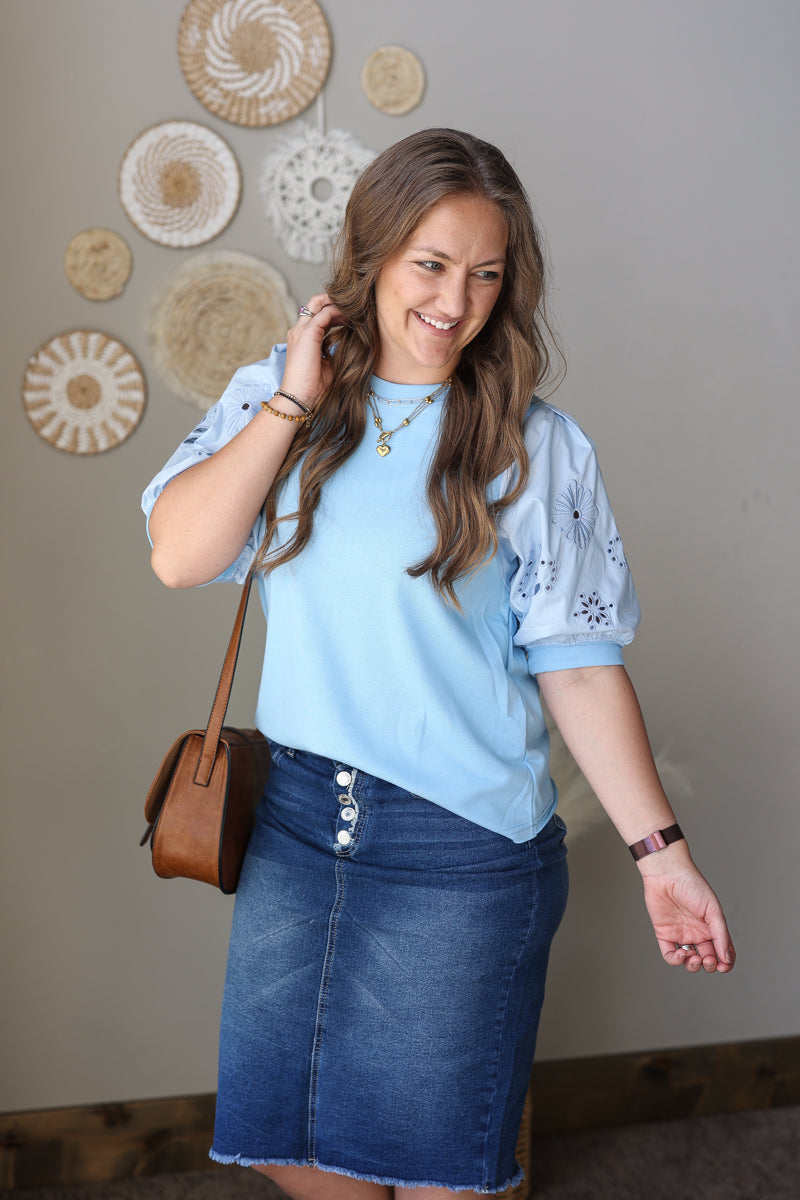 Woman wearing a light blue top and denim skirt with a brown handbag indoors.