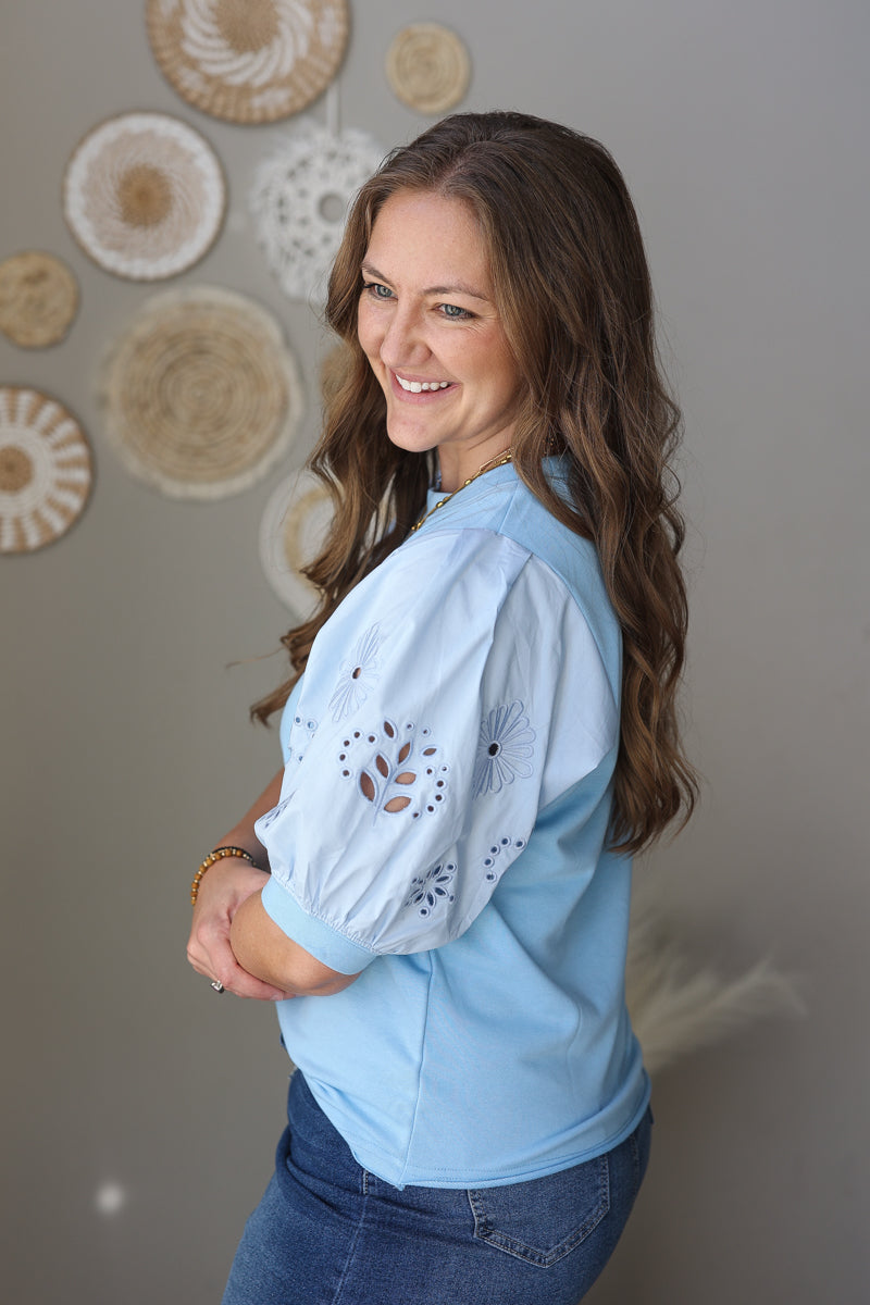 Woman wearing a light blue blouse with lace details, standing against a neutral wall with decorative items.
