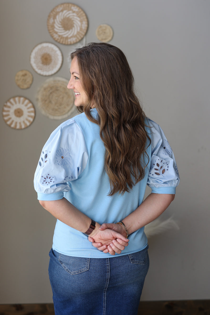 Woman wearing a light blue blouse with lace sleeves against a neutral wall.