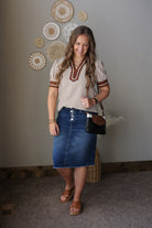Woman wearing a striped shirt and denim skirt standing indoors with decorative wall art in the background.