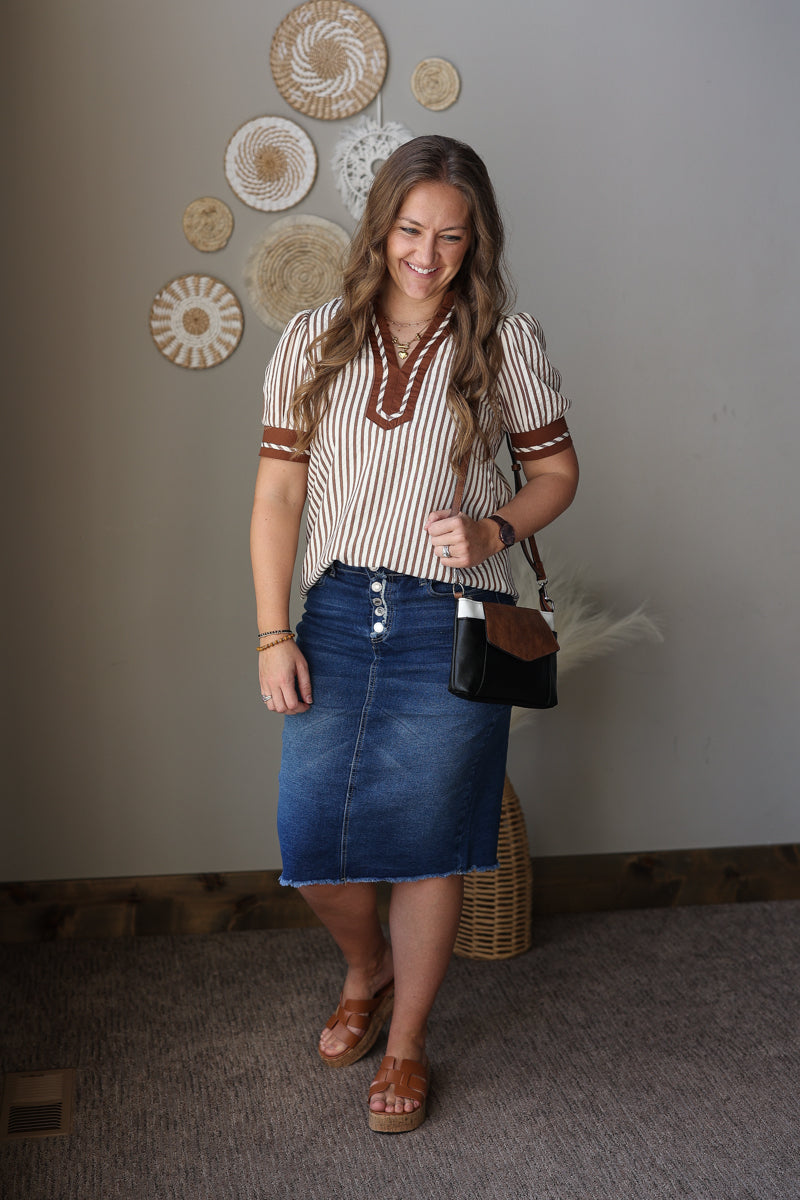 Woman wearing a striped shirt and denim skirt standing indoors with decorative wall art in the background.