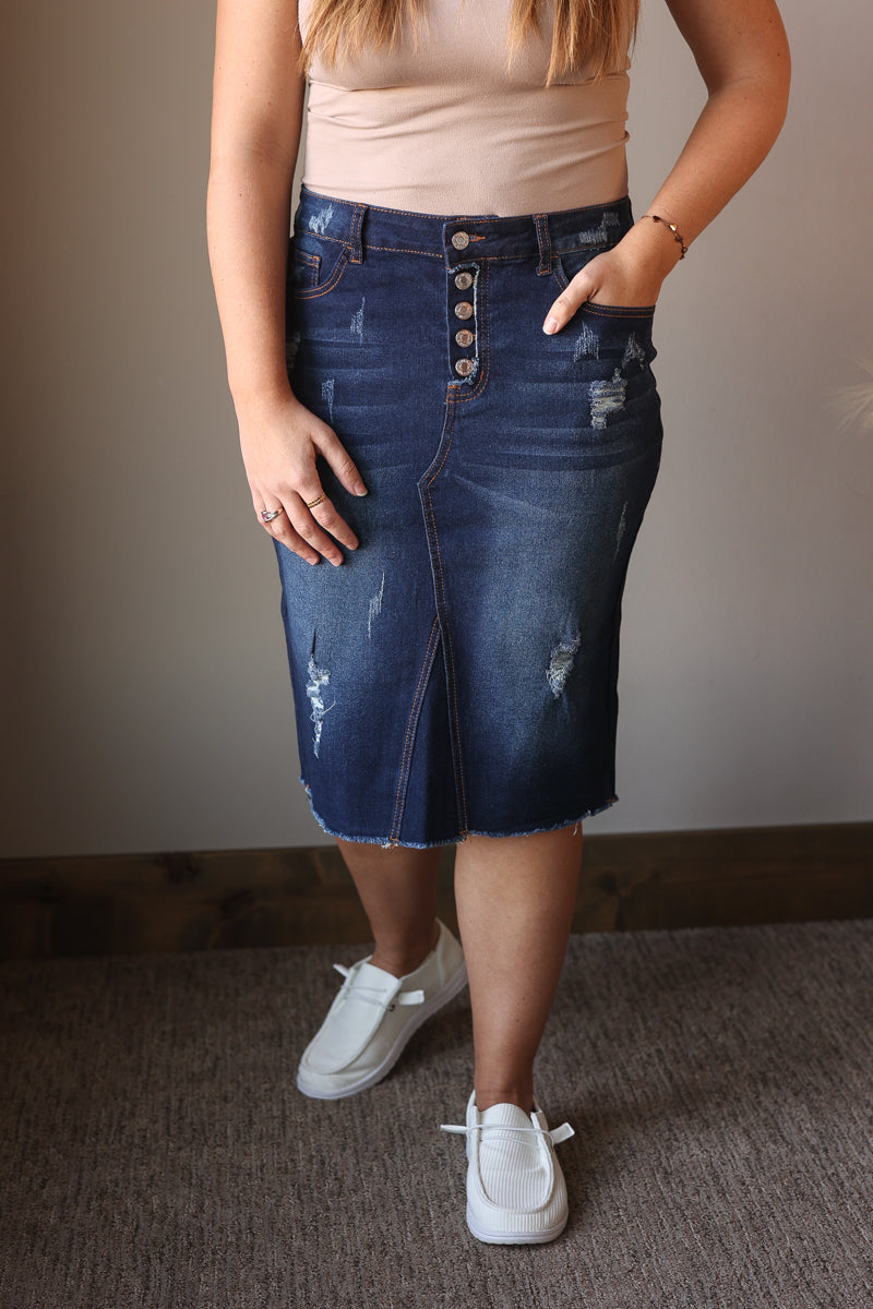 Person wearing a blue denim skirt with button details, beige top, and white sneakers indoors.