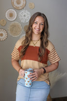 Woman wearing a tan colorblock top holding a mug with decorative elements on the wall behind her