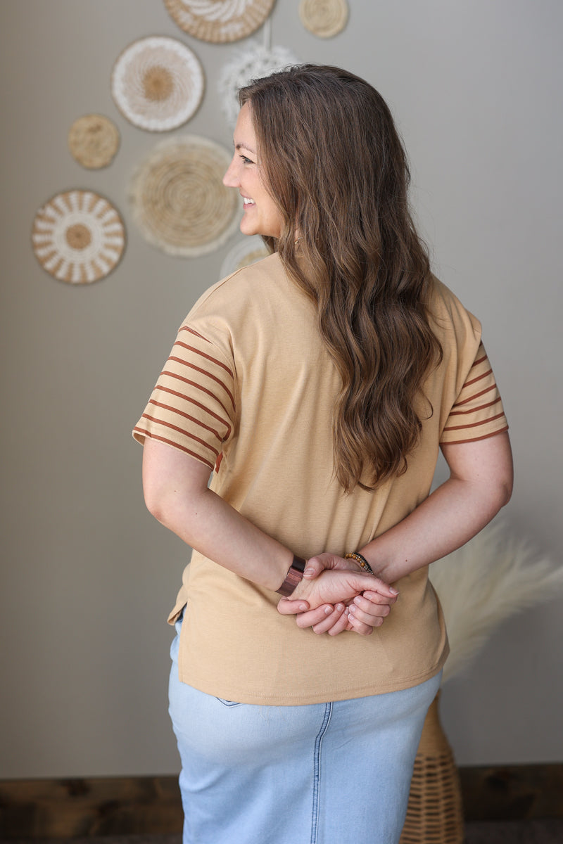 Woman wearing a beige top with striped sleeves and light blue jean skirt, standing in a room with decorative wall art.