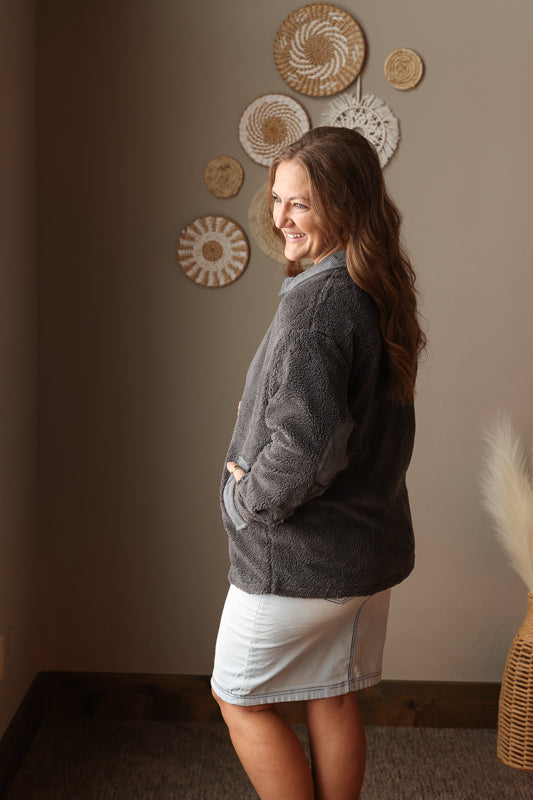 Woman wearing a gray fleece jacket and light wash denim skirt standing in a room with decorative wall plates.