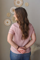 Woman wearing a pink textured top and blue jean skirt standing against a neutral wall with decorative elements.