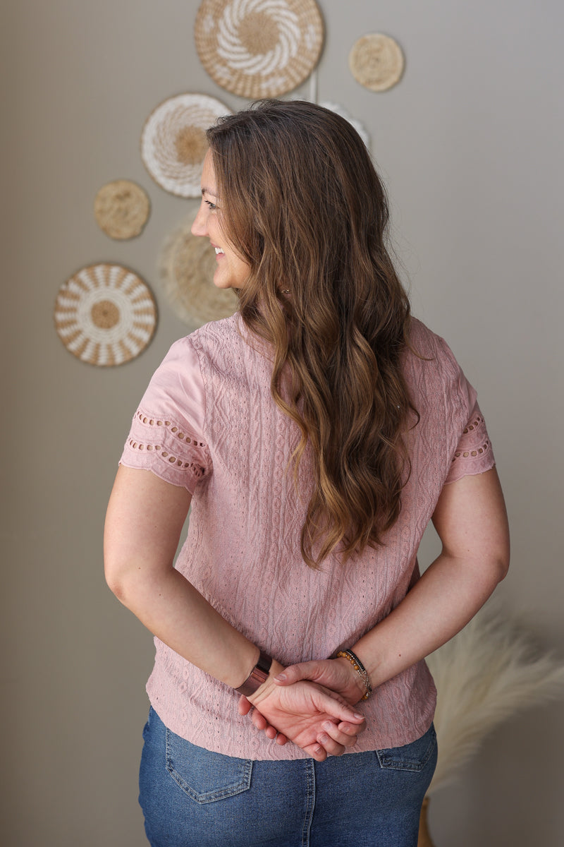 Woman wearing a pink textured top and blue jean skirt standing against a neutral wall with decorative elements.