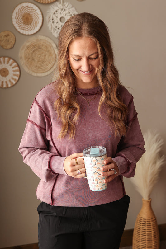 Woman wearing Burgundy Mineral Wash Side Slit Sweatshirt with relaxed fit, styled casually with joggers, perfect for comfortable everyday wear. Woman holding a mug with decorative elements on a neutral wall background