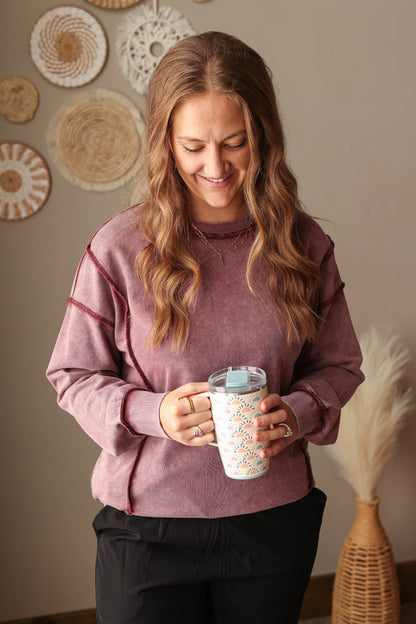 Woman wearing Burgundy Mineral Wash Side Slit Sweatshirt with relaxed fit, styled casually with joggers, perfect for comfortable everyday wear. Woman holding a mug with decorative elements on a neutral wall background