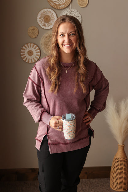 Woman holding a mug in a room with decorative wall art and a vase. Woman wearing Burgundy Mineral Wash Side Slit Sweatshirt with relaxed fit, styled casually with joggers, perfect for comfortable everyday wear