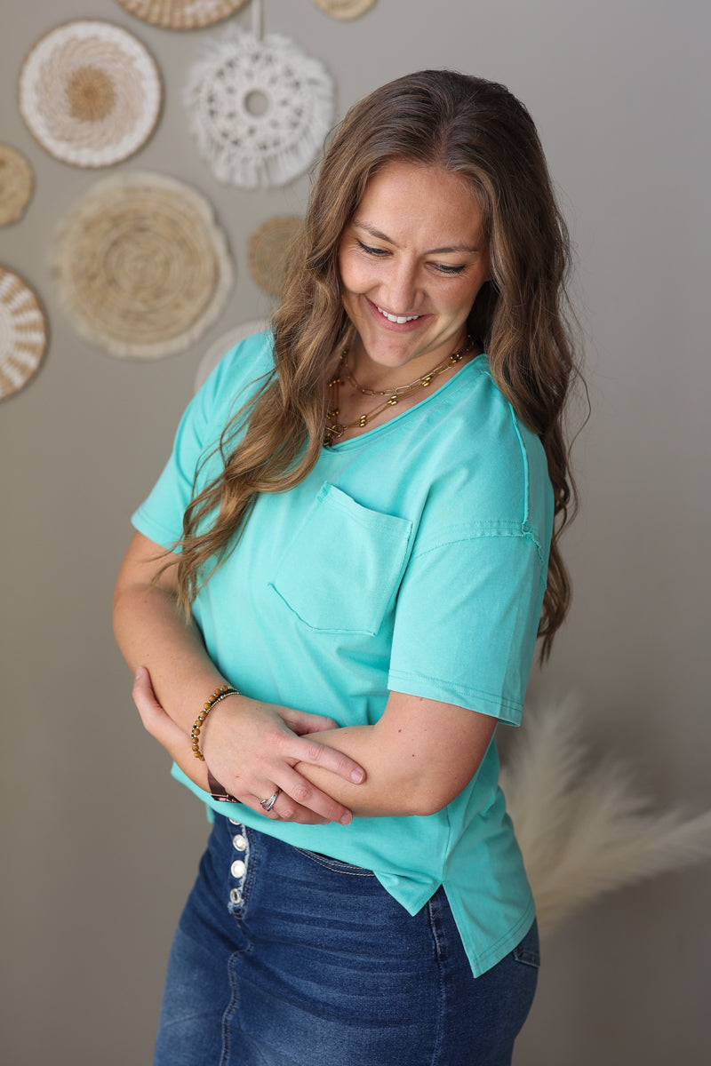Woman wearing a turquoise shirt and blue jean skirt against a neutral wall with decorative elements.