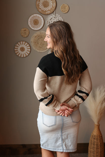 Woman wearing a black and beige sweater with a light wash denim skirt, standing in a room with decorative wall art.
