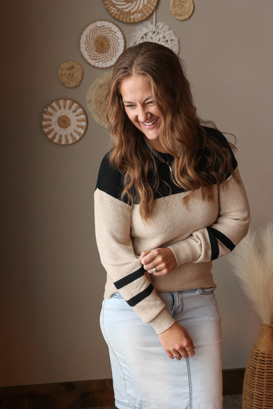 Woman wearing a beige sweater with black sleeves in a room with decorative wall art.