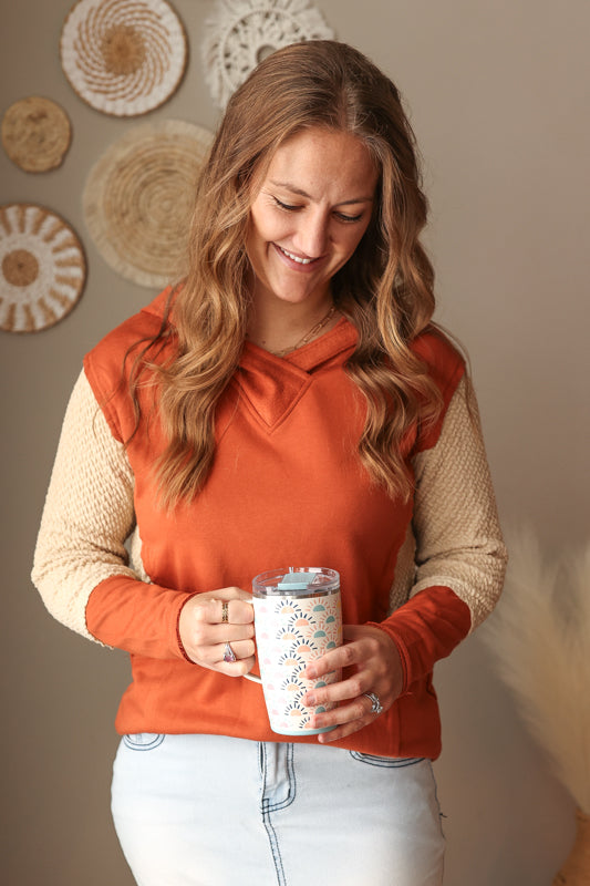Woman in an orange hoodie holding a mug with decorative elements on the wall behind her.