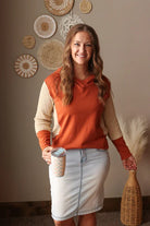Woman in a rust-colored sweater and light wash knee length skirt holding a coffee cup, standing against a wall with decorative baskets.