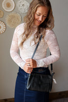 A woman wearing a white floral lace long-sleeve top and a dark blue denim skirt, carrying a black leather crossbody bag. She is standing indoors against a neutral gray wall decorated with circular woven wall art.