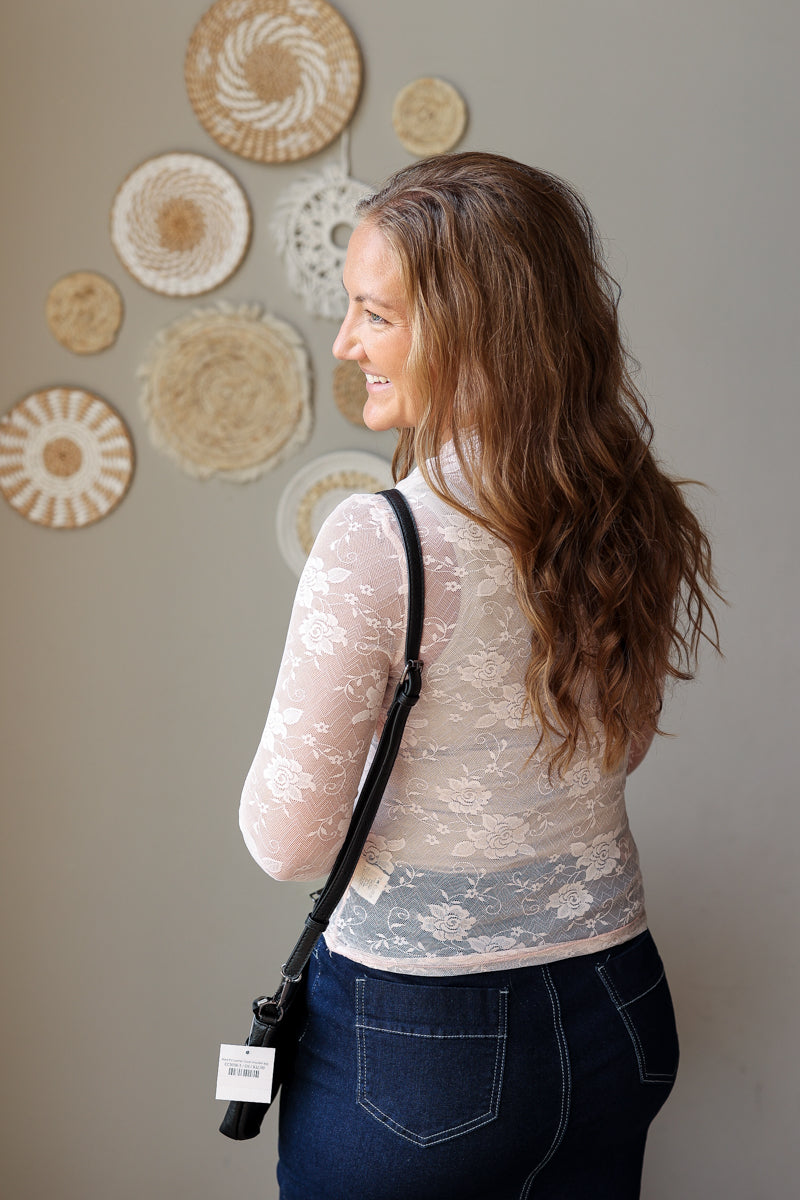 A woman with long brown hair is shown from the back and side, wearing a sheer beige lace long-sleeve top and dark blue jean skirt, carrying a black shoulder bag. She is standing against a gray wall decorated with circular woven wall art.