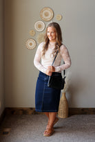 A woman posing in a cream lace long-sleeve mock neck top and a dark blue denim midi skirt, carrying a black crossbody bag. She is standing in a room with a neutral gray wall decorated with circular woven wall art.