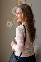 A woman with long brown hair is shown from the side and back, wearing a beige sheer lace long-sleeve top and dark blue jean skirt, carrying a black shoulder bag against a neutral wall with decorative circular wall art.