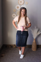 Woman holding a colorful mug in a room with decorative wall art and a vase, wearing a khaki crewneck tee and black denim midi skirt for spring summer outfits that are modest for women.