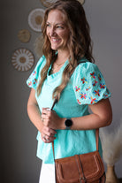 Woman wearing a turquoise top with floral sleeves, holding a brown bag indoors.