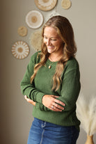 A woman with long light brown hair smiling and looking down, wearing a green waffle-knit sweater and blue jean skirt, standing in front of a neutral wall decorated with circular woven wall art.