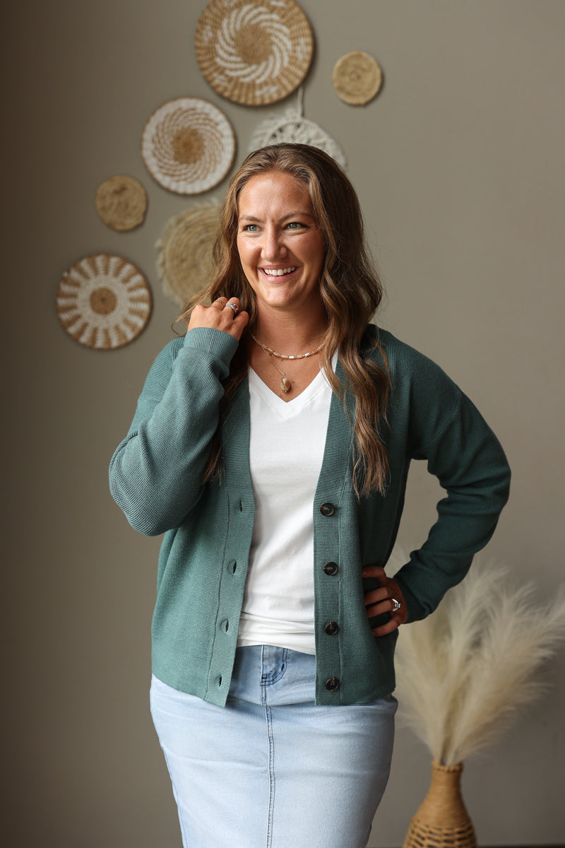 A woman smiling and posing in a sage green button-up cardigan over a white V-neck top and light blue jean skirt, standing against a warm gray wall decorated with circular woven wall art.