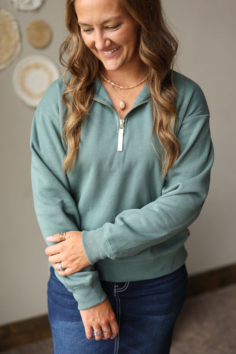A woman wearing a sage green quarter-zip sweatshirt and dark blue jean skirt, standing indoors against a neutral wall.