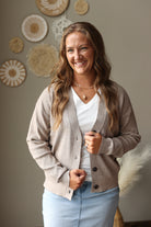 A smiling woman with long wavy light brown hair stands indoors wearing a light taupe knit cardigan over a white V-neck top and light blue jean skirt. The background is a warm neutral wall with decorative woven wall art and a pampas grass accent.