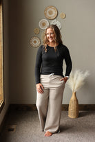 A woman stands indoors wearing a black long-sleeve top and light beige wide-leg drawstring pants. The room has a warm neutral wall with decorative woven wall art, a carpeted floor, and a woven vase with pampas grass.