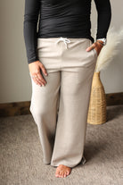 A person wearing beige wide-leg lounge pants with a white drawstring, paired with a black long-sleeve top, standing barefoot on a textured carpet.
