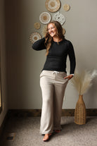 A woman stands indoors wearing a fitted black long-sleeve top and light beige drawstring pants. The room has a warm neutral wall with decorative woven wall art, carpet, and a wicker vase with dried pampas grass.