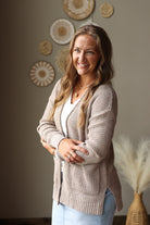 A smiling woman with long light brown hair stands indoors wearing a beige waffle-knit cardigan over a white top and light blue jean skirt. The background shows a warm neutral wall with decorative woven wall art and a vase with dried pampas grass.