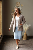 A woman stands indoors wearing a light brown waffle-knit cardigan over a white top, a light blue midi skirt, and cream sneakers. The background features a warm gray wall with decorative circular wall art and a woven vase with dried pampas grass.