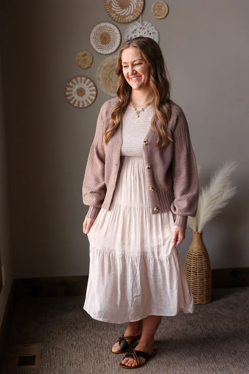 Woman wearing a light pink cardigan over a white dress, standing in a room with decorative wall art.