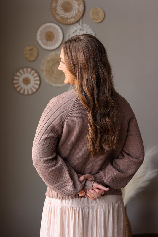 Woman wearing a brown sweater and beige skirt standing against a wall with decorative plates.
