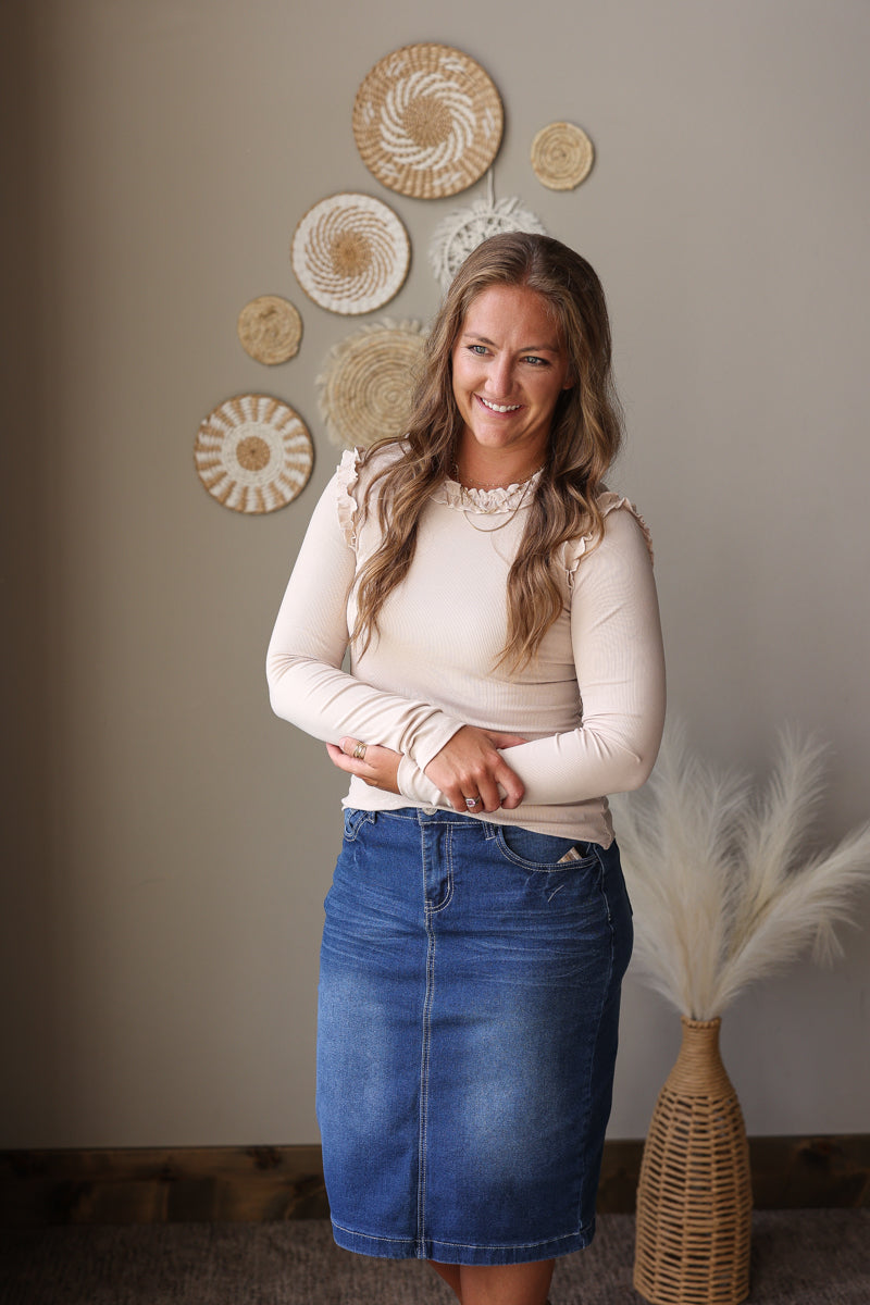 A woman stands indoors wearing a fitted cream long-sleeve top and a blue denim skirt. She is posed in front of a beige wall with woven circular wall decor and a vase of pampas grass.